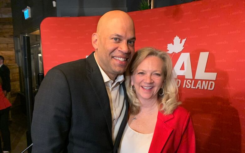 Man in dark-grey suit stands in front of Liberal sign with woman in red suit. 