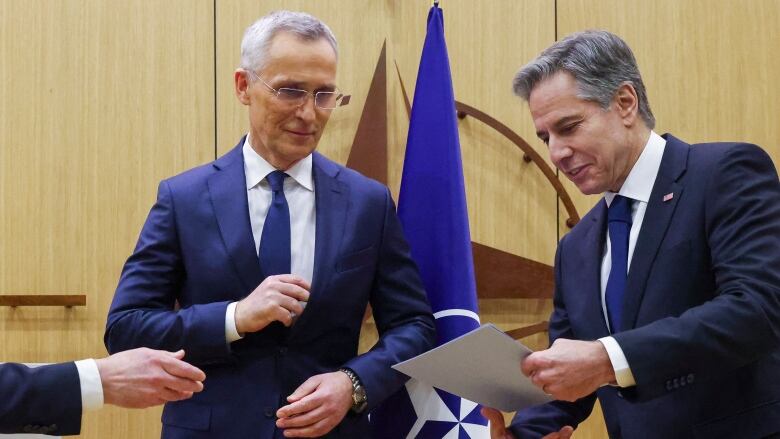 Two men in dark suits are shown standing, with one holding documents.