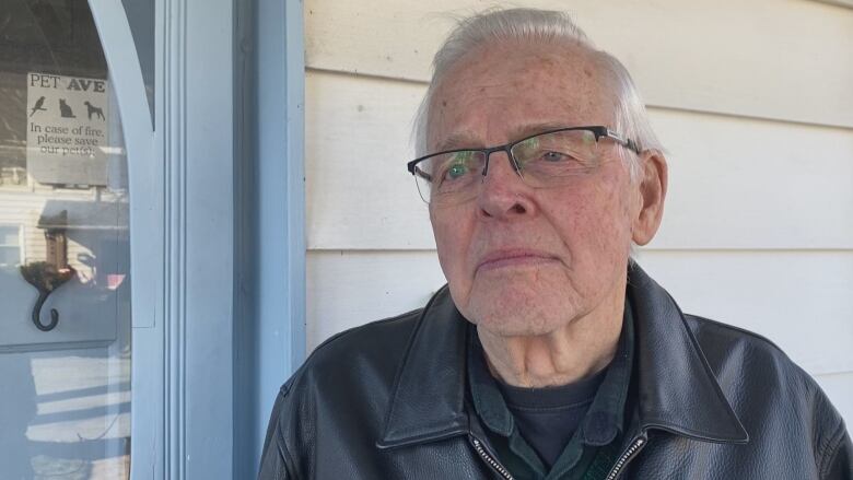 A man with short white hair and glasses wears a leather jacket and navy blue shirt. He stands in front of a doorway.