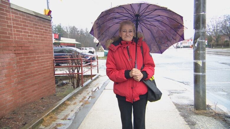 A women in a red coat with an umbrella stands on the sidewalk.