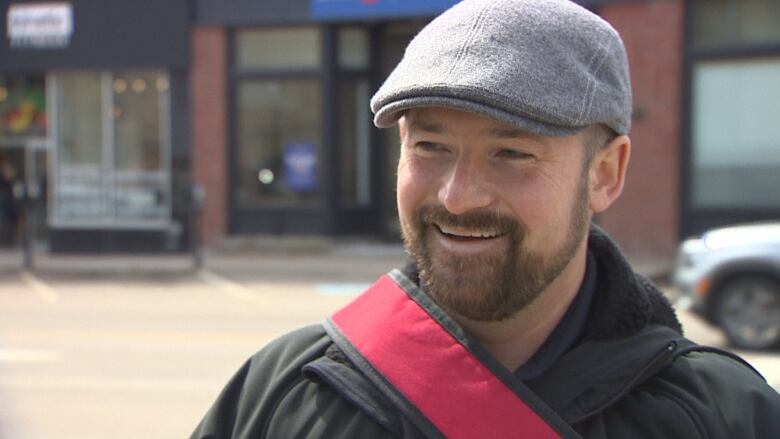 A man stands on the street wearing a flat cap and smiling. 