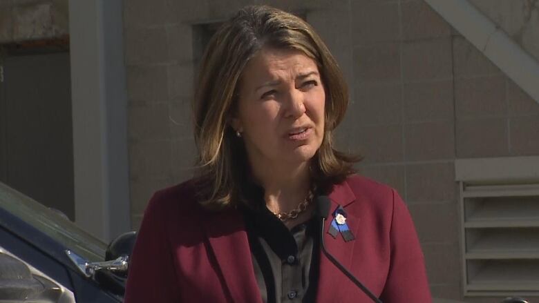 Smith wears a burgundy jacket with a blue pin as she squints in the sunlight speaking to reporters. 