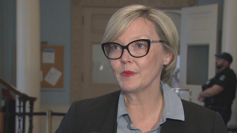 A woman with blonde hair and glasses wears a black blazer with a grey top. She is standing near a staircase.