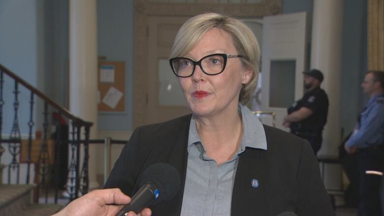 A woman with blonde hair and glasses wears a black blazer with a grey top. She is standing near a staircase.