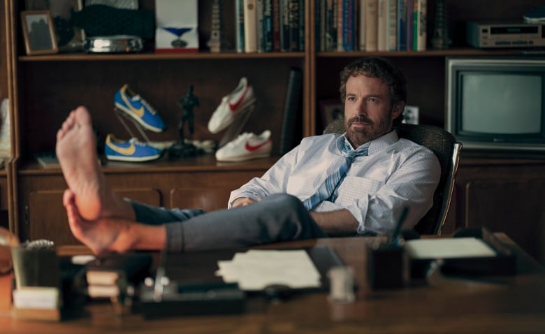 A businessman with a beard leans back in his chair and puts his bare feet on his desk. 