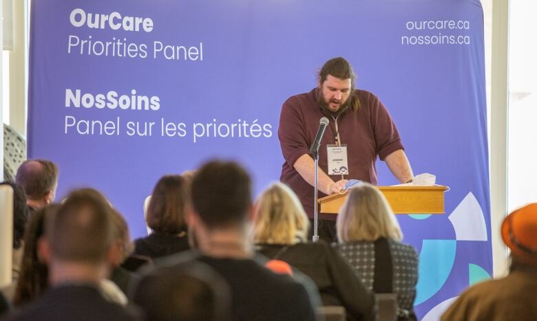 Joshua McCoy stands at a podium, with a banner saying 'OurCare Priorities Panel' behind him. 