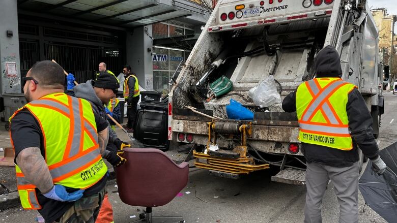 A man in a yellow safety best puts his hand on an office chair on rollers near other men in yellow safety vests congregating near a garbage truck where they are preparing to throw refuse and other items.