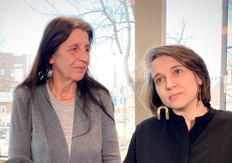 Two women stand in front of a window and smile.