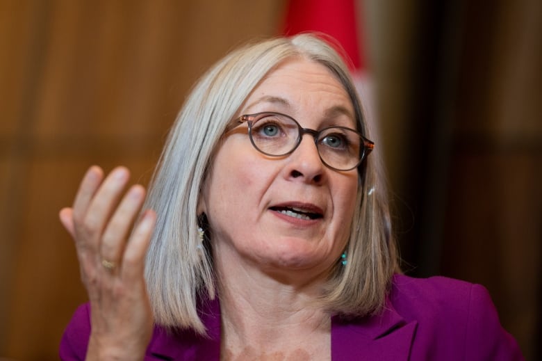 A woman with shoulder-length white hair and glasses, wearing a purple jacket, gestures as she speaks to reporters.