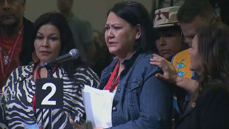 A woman speaks into a microphone as a group of people surround her to show their support.