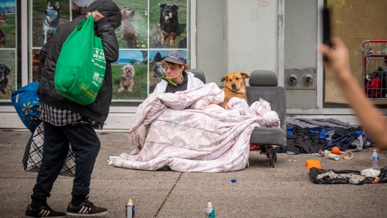 A man and his dog sit on a bench seat from a vehicle under a quilt as a person walks by carrying bags