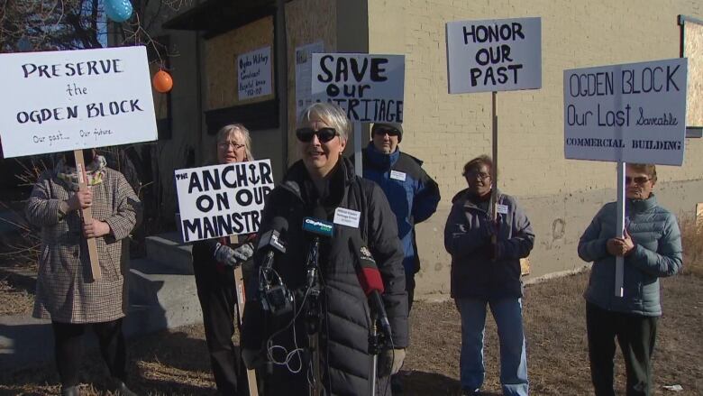 A woman in a black puffer jacket stands in front of microphones. Behind her, five people stand holding signs. 