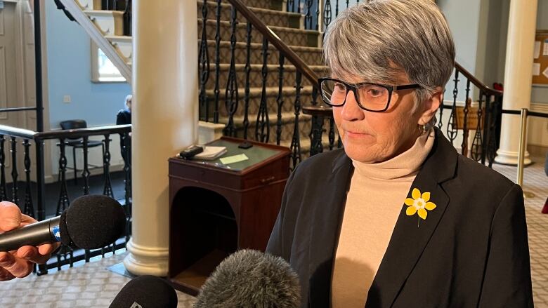 A woman with white hair and glasses stands in front of microphones.