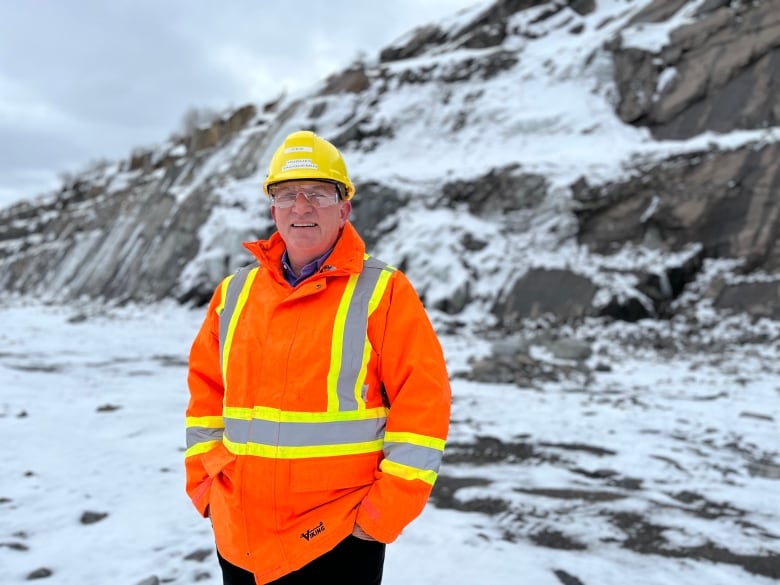 A man wearing a hardhat and orange jacket poses in front of a rock face.