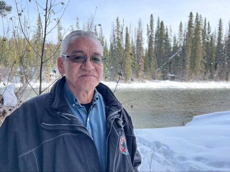 An older man wearing glasses, a denim shirt and a blue jacket stands next to a snowy riverbank.