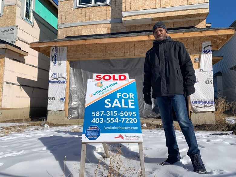 A man wearing jeans and a jacket stands on a snowy yard, beside a sign that says 