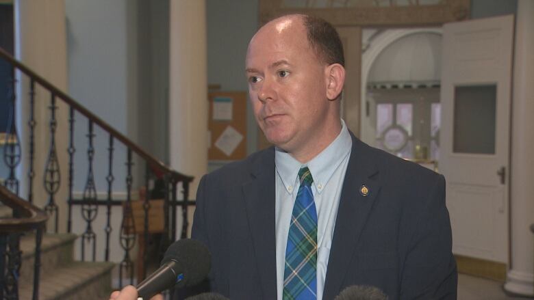 A man, wearing a suit with a blue and green tartan tie, stands in front of microphones.