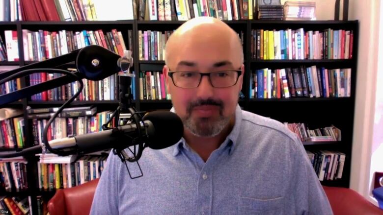 A man of south Asian descent stands in front of a bookshelf, with a microphone in front of him.