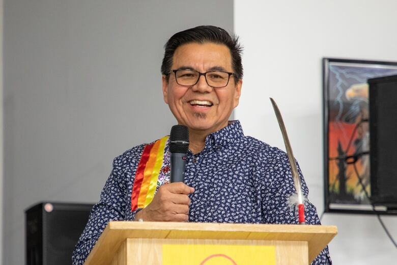 A man has short black hair, eyeglasses and wears a floral shirt with red, orange and yellow stripes down the right side. He stands at a podium speaking into a microphone held in his hand.