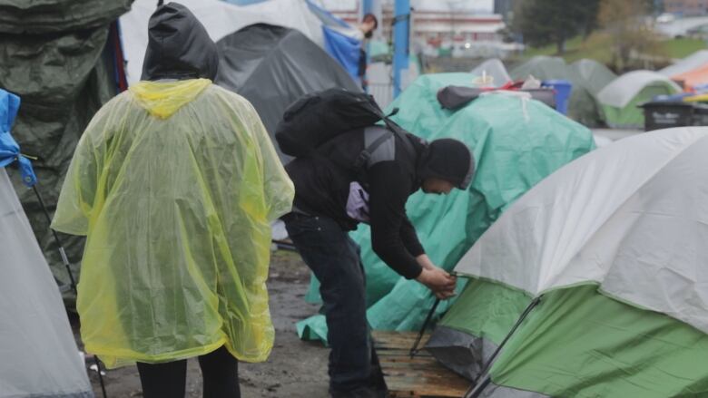 A man sets up a tent while another man in a yellow poncho watches.