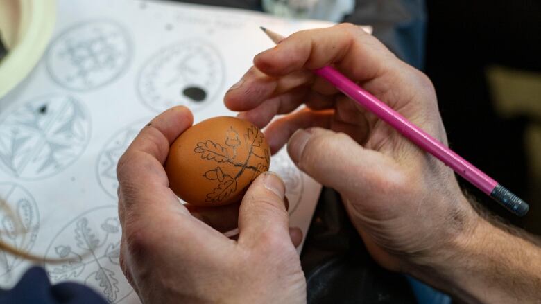 Hands hold an egg with a design drawn in pencil on it.