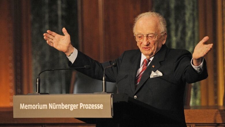 A elderly person gestures while speaking at a lectern.