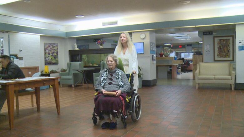 A woman pushes an elderly woman in a wheelchair in a long-term care home. 