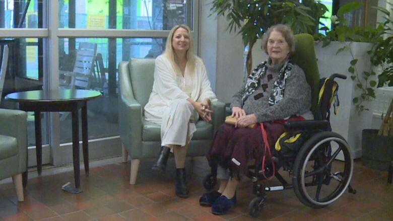 A woman sits on a green armchair, beside an elderly woman sitting in a wheelchair. 