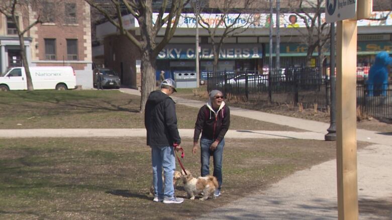 People stand in a park, one is laughing and the other has two small dogs on a leash.