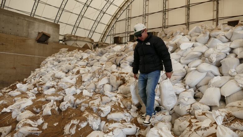 A young man wearing sunglasses and a baseball cap carries two sandbags as he descends a large pile of sandbags.