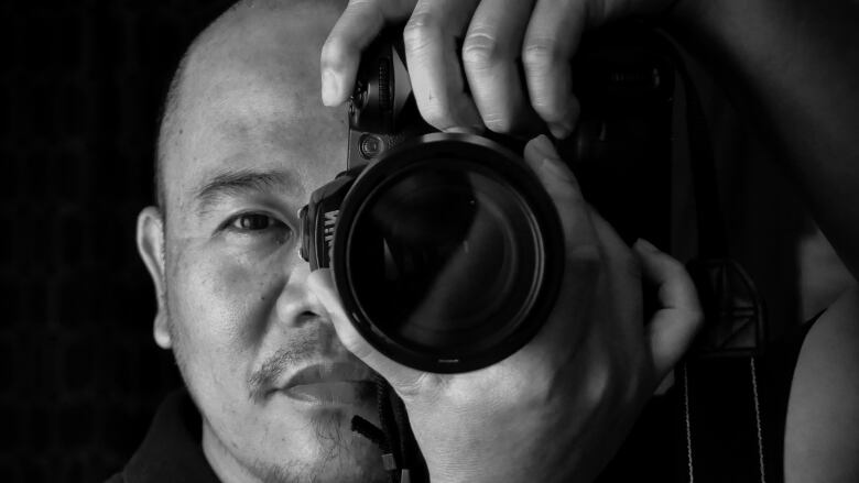 Man with bald head holds and points a camera at the viewer. He is wearing a black t-shirt. 