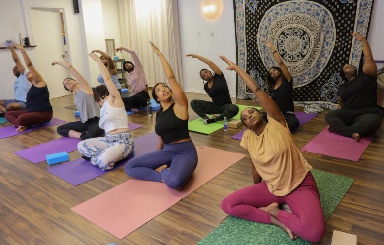 About a dozen people sit on multi-coloured mats in a yoga studio and stretch their arms above their heads. 