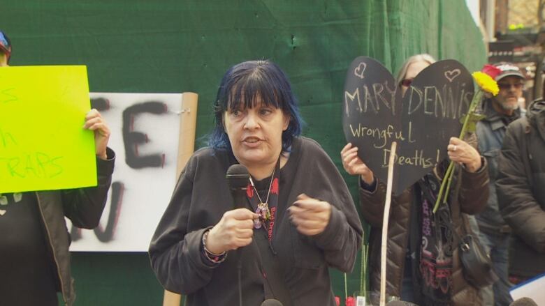 A woman holds a microphone at a rally, with other people visible behind her. One of them is holding up a heart-shaped sign that reads 'Mary Dennis Wrongful Deaths'.