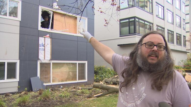 A man with a beard and long flowing hair points to a second-storey window that is being replaced as tree logs are visible behind him.