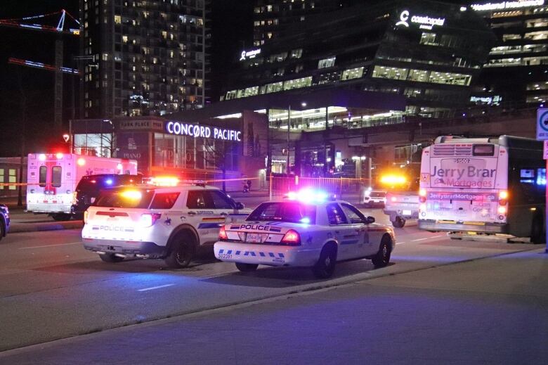 Flashing police cars sit parked behind a bus, with ambulances and SkyTrain tracks visible in the background.