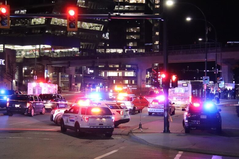 Flashing police cars surround a taped-off intersection, with a bus and SkyTrain tracks visible in the background.