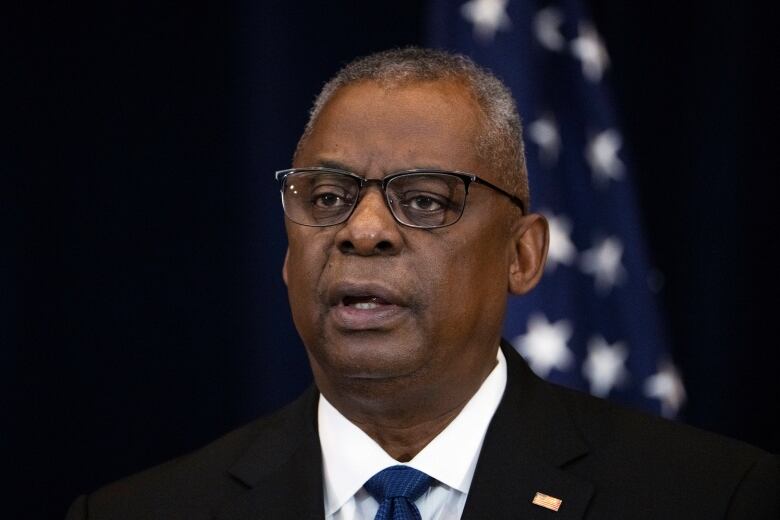 A bespectacled man in a suit and tie with a pin on his jacket on his lapel is shown in closeup speaking.