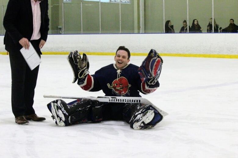 David Lewis, wearing hockey goaltending gear, is sitting on the ice with his goalie stick lying across his lap. He is smiling.