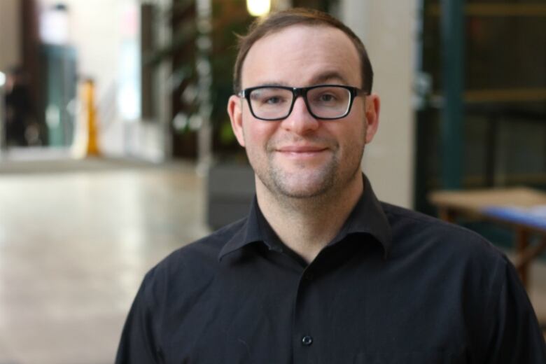 A man with glasses and a black shirt stands in a hallway.