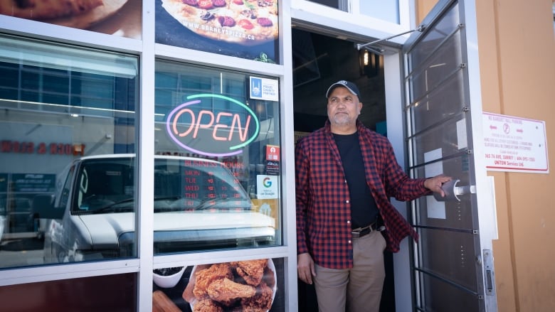 A man holds a restaurant door open and is looking outside. He is wearing a hat with a red and black checkered shirt. 