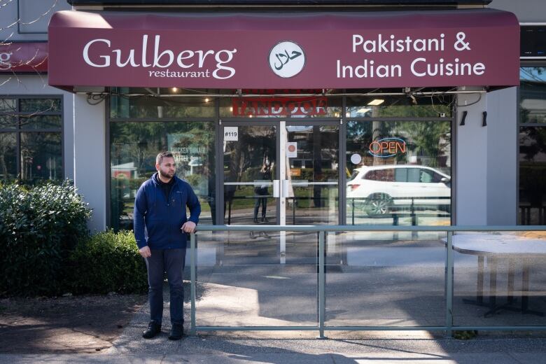 A man in a blue hoodie stands in front of a restaurant with a sign a sign saying 'Gulberg Restaurant, Pakistani & Indian Cuisine.'