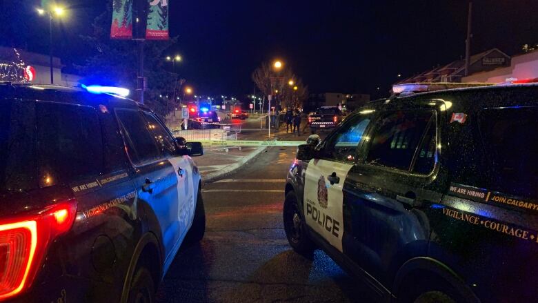 A Calgary intersection at night seen between two police cars with police crime scene yellow tape in the distance.