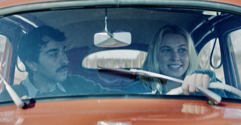 A young man and women are shown through the front windshield of a car.