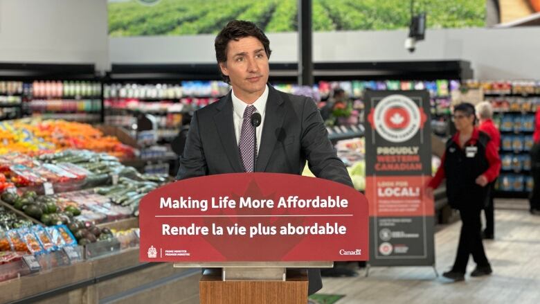 A man in a black suit with a purple tie speaks stands at a podium with the phrase 