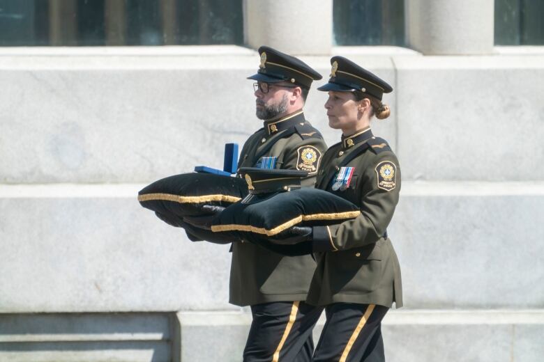 Two officers in brown carry velvet pillows with a peaked cap and medal box.