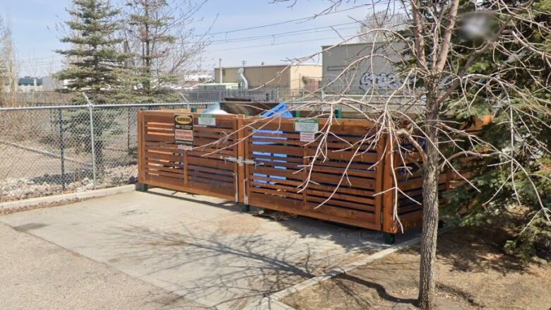 A small wooden garbage containment structure in the corner of a parking lot in an industrial area.