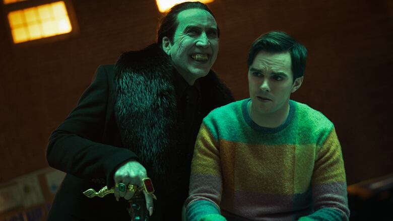 A smiling, pallid man with pointed teeth leans over a grimacing younger man wearing a colourful sweater. The pale man wears all black, has a number of large rings on his hand and is leaning on an ornate cane. 