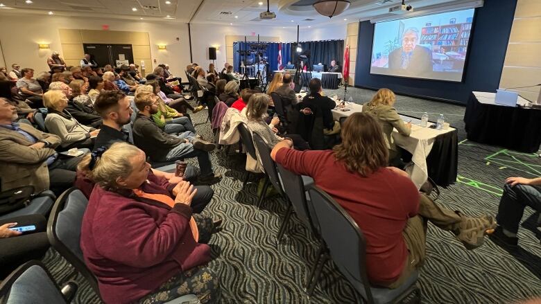 A crowd of people sitting in chairs watch as a man gives a presentation on a large projector screen