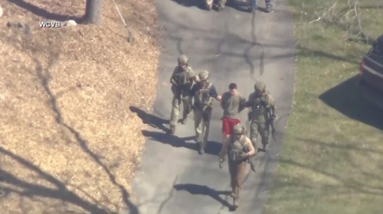 A video still from an aerial view shows a man in shorts and a tshirt with hands behind his back being escorted by four people in military fatigues.