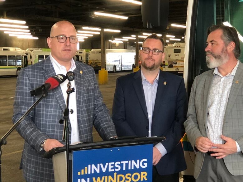 A man stands behind a podium in a warehouse, flanked by two other men. 